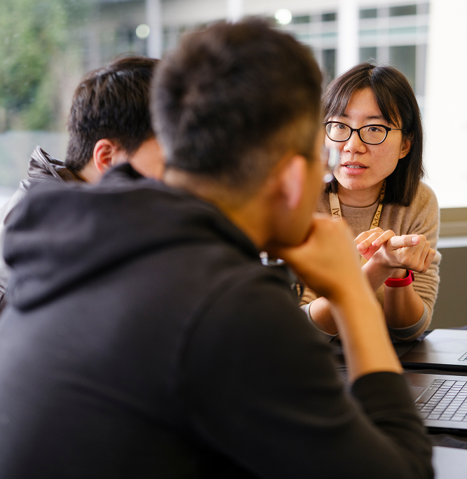 A woman wearing glasses speaks and gestures with her hands while sitting at a table with two people, who have their backs to the camera, in a bright indoor setting.