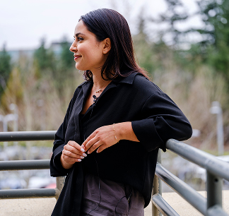A woman with dark hair, wearing a black top, stands outdoors leaning on a railing, looking to the side and smiling. Trees and a blurred background are visible behind her.