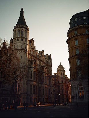 A city street at sunset with historic, ornate buildings featuring towers and domes; golden sunlight casts long shadows, and a few pedestrians and cars are visible on the quiet road.