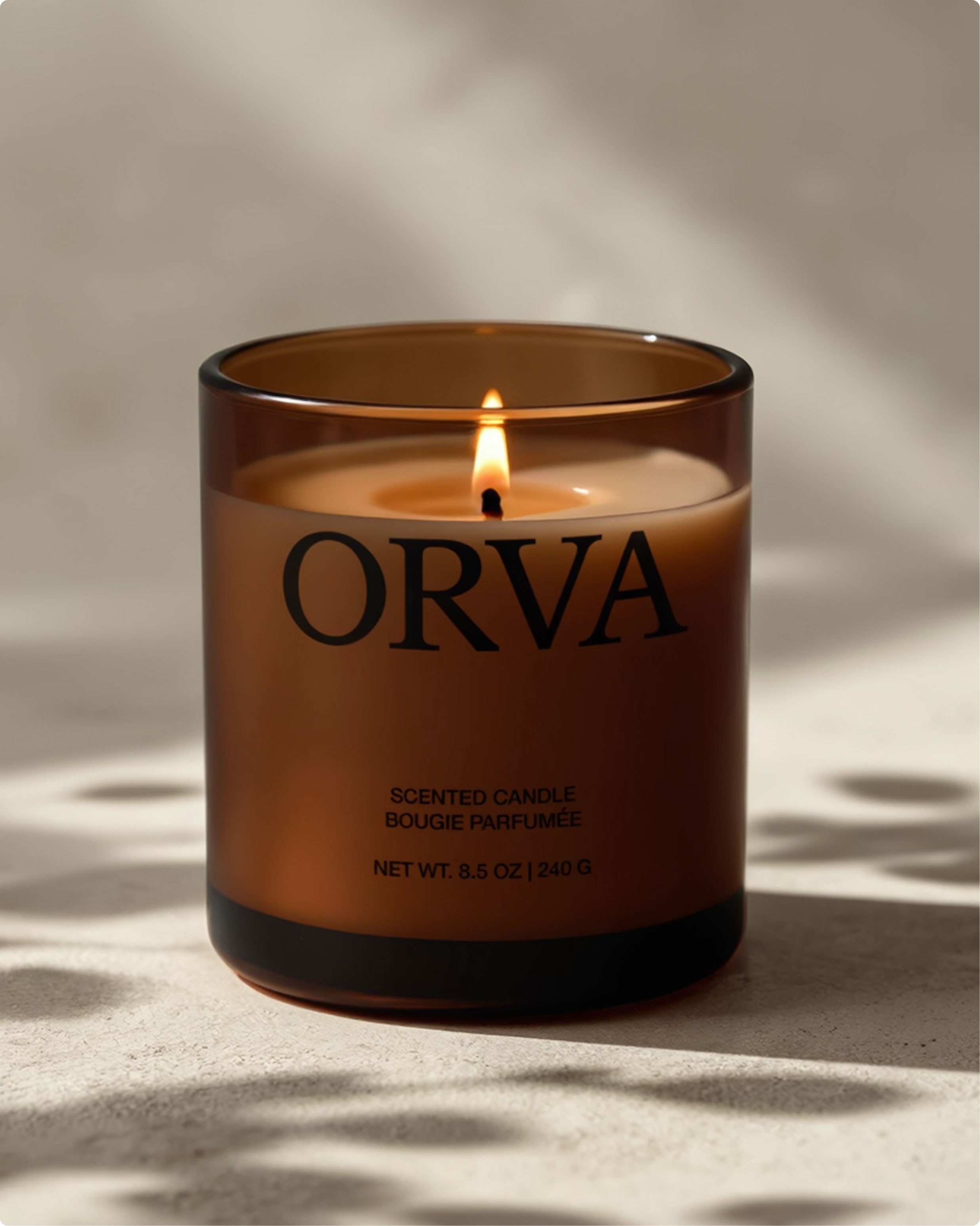 A brown glass candle labeled "ORVA" is lit, with a single flame and soft shadows cast on a neutral surface. The label indicates it is a scented candle, weighing 8.5 oz or 240 g.