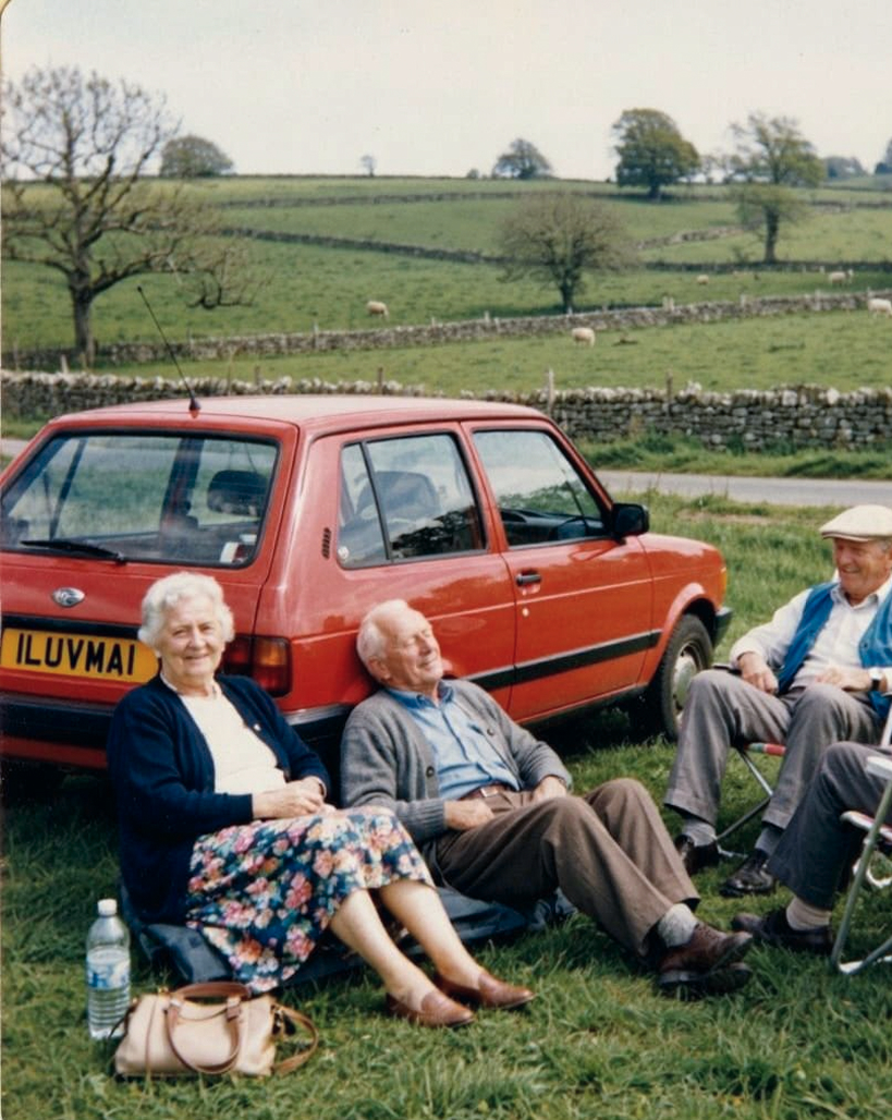 Three elderly people relax on the grass in front of a red car with the license plate "ILUVMAl," enjoying a countryside setting with green fields and stone walls in the background.