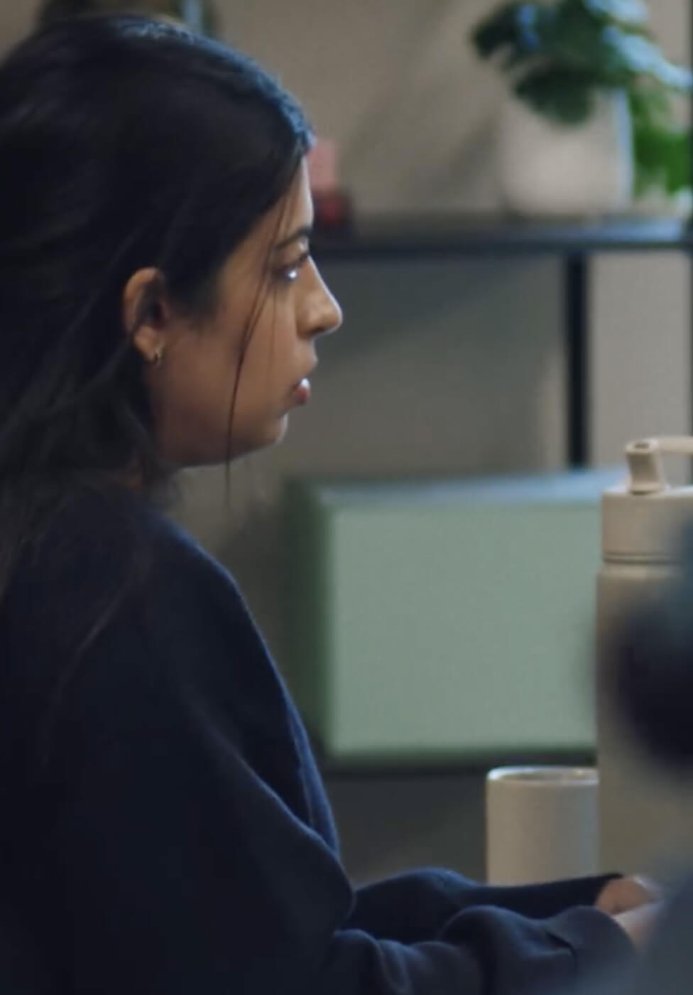 A woman with long dark hair, wearing a dark sweater, sits at a desk with a large water bottle and a mug, looking intently at something off-camera. Blurred shelves and a plant are in the background.