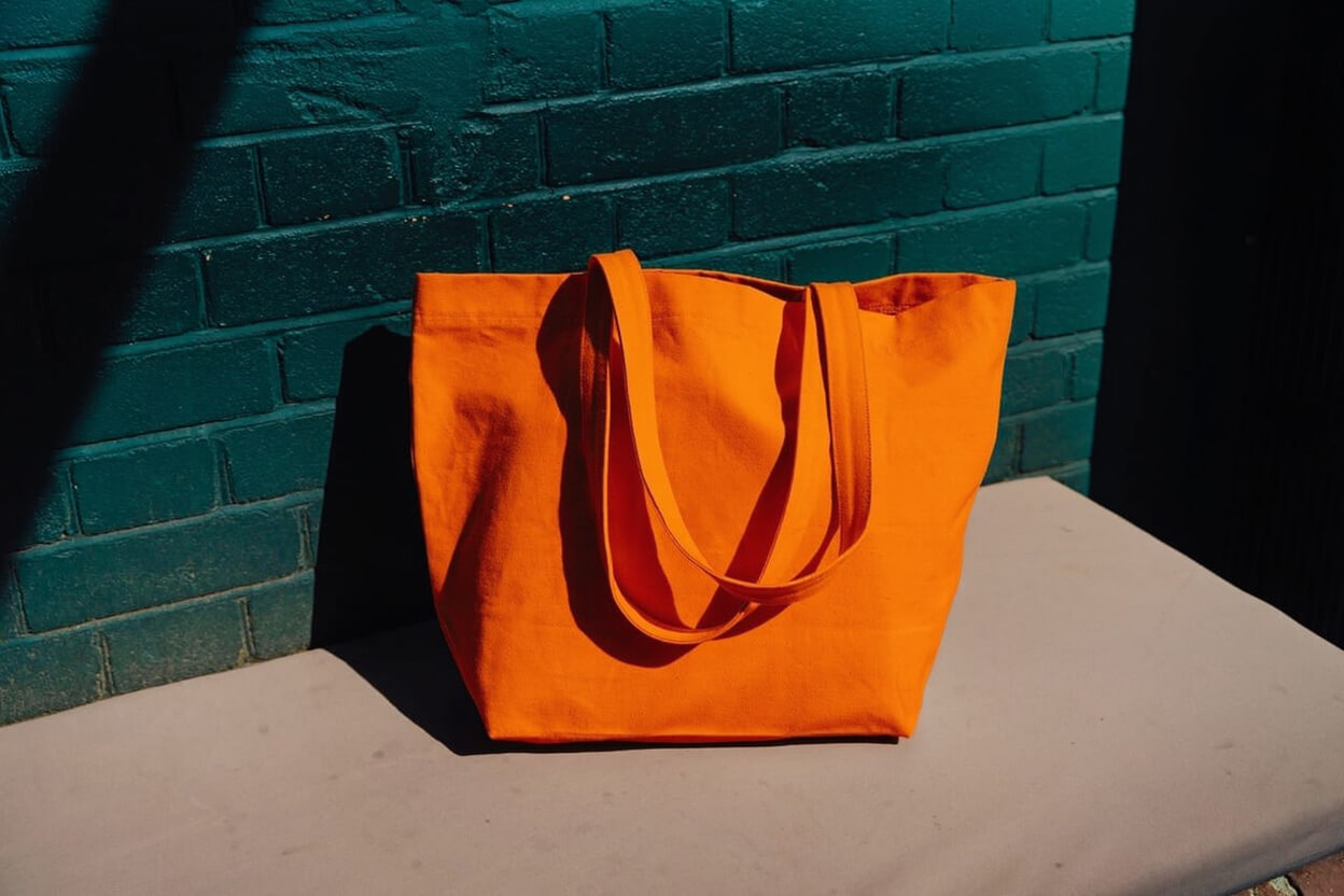 A bright orange tote bag with two handles sits on a light-colored surface in front of a teal brick wall, partially illuminated by sunlight.