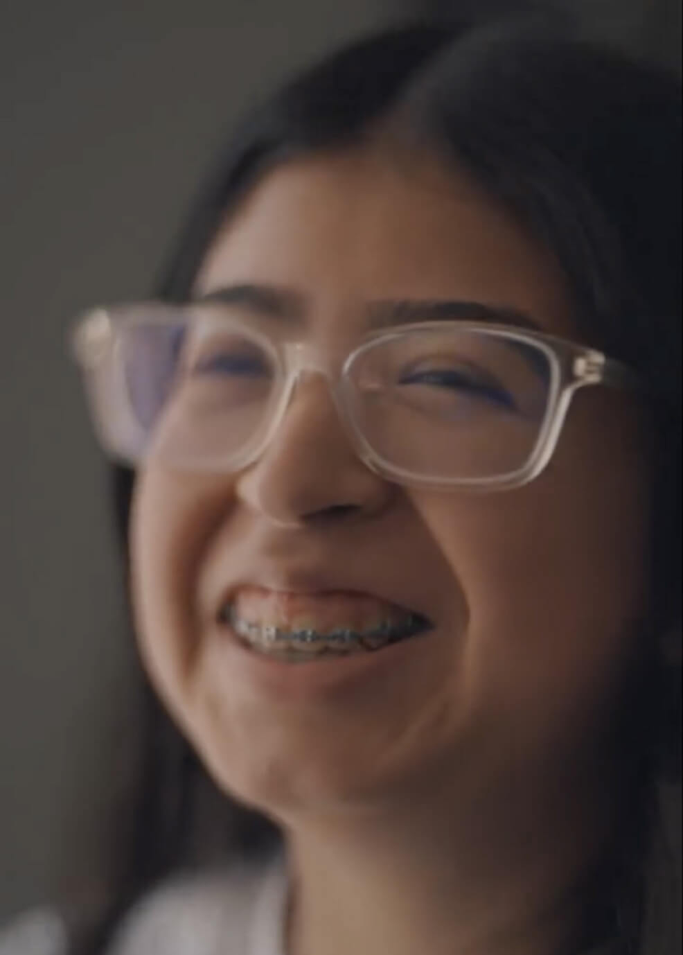 A young person with long dark hair, wearing clear-framed glasses and braces, smiles brightly against a blurred background.