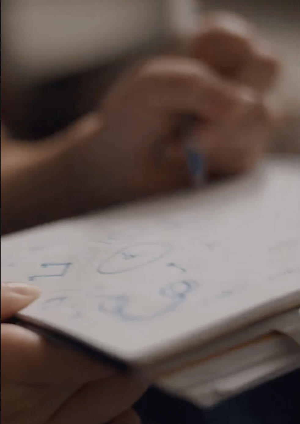 A close-up of a person sketching abstract shapes and diagrams with a blue pen on a white notepad. The image is slightly blurred, focusing on the hand and the drawing.