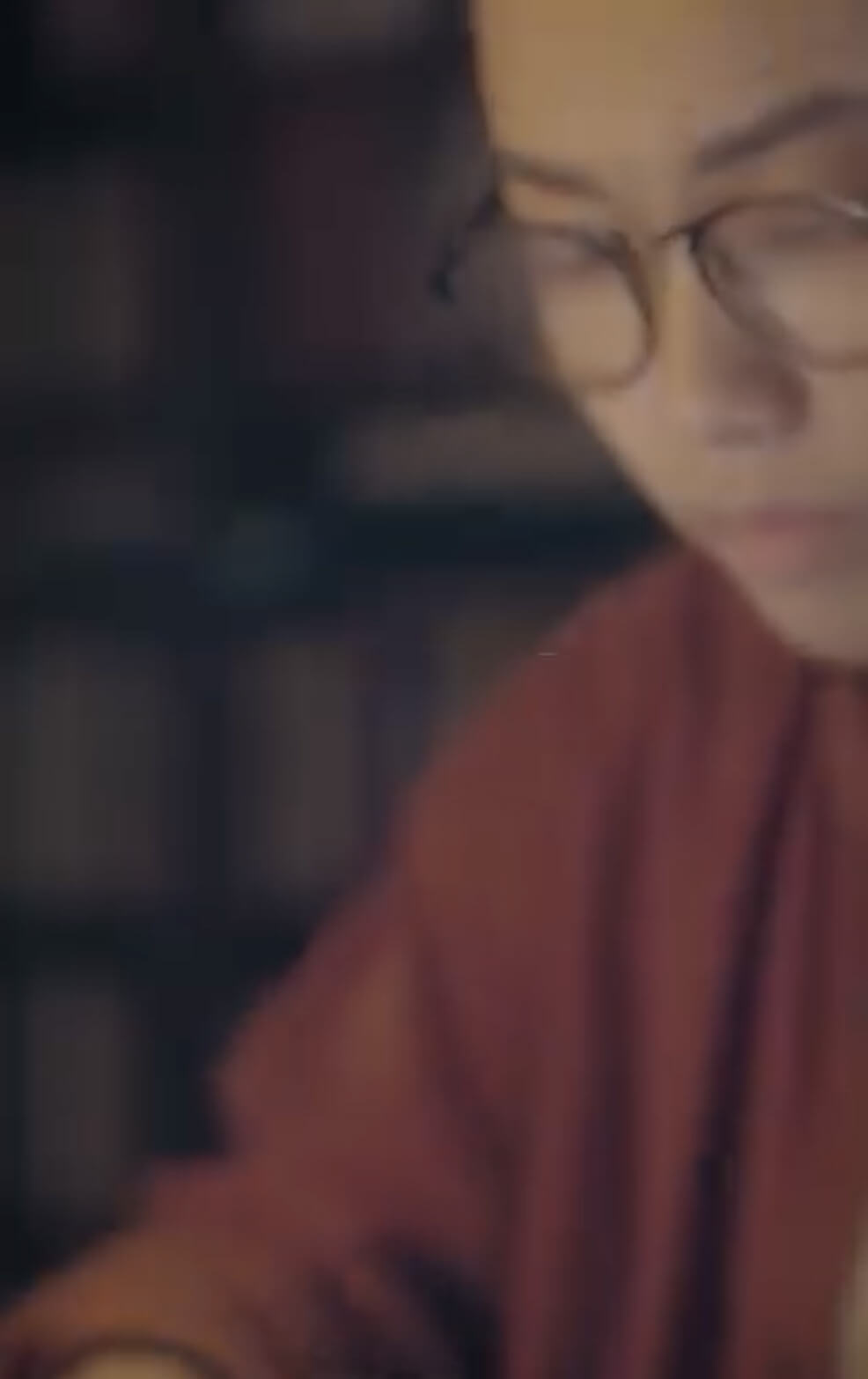 A person wearing glasses and an orange sweater appears to be focused on something in front of them, with bookshelves blurred in the background.