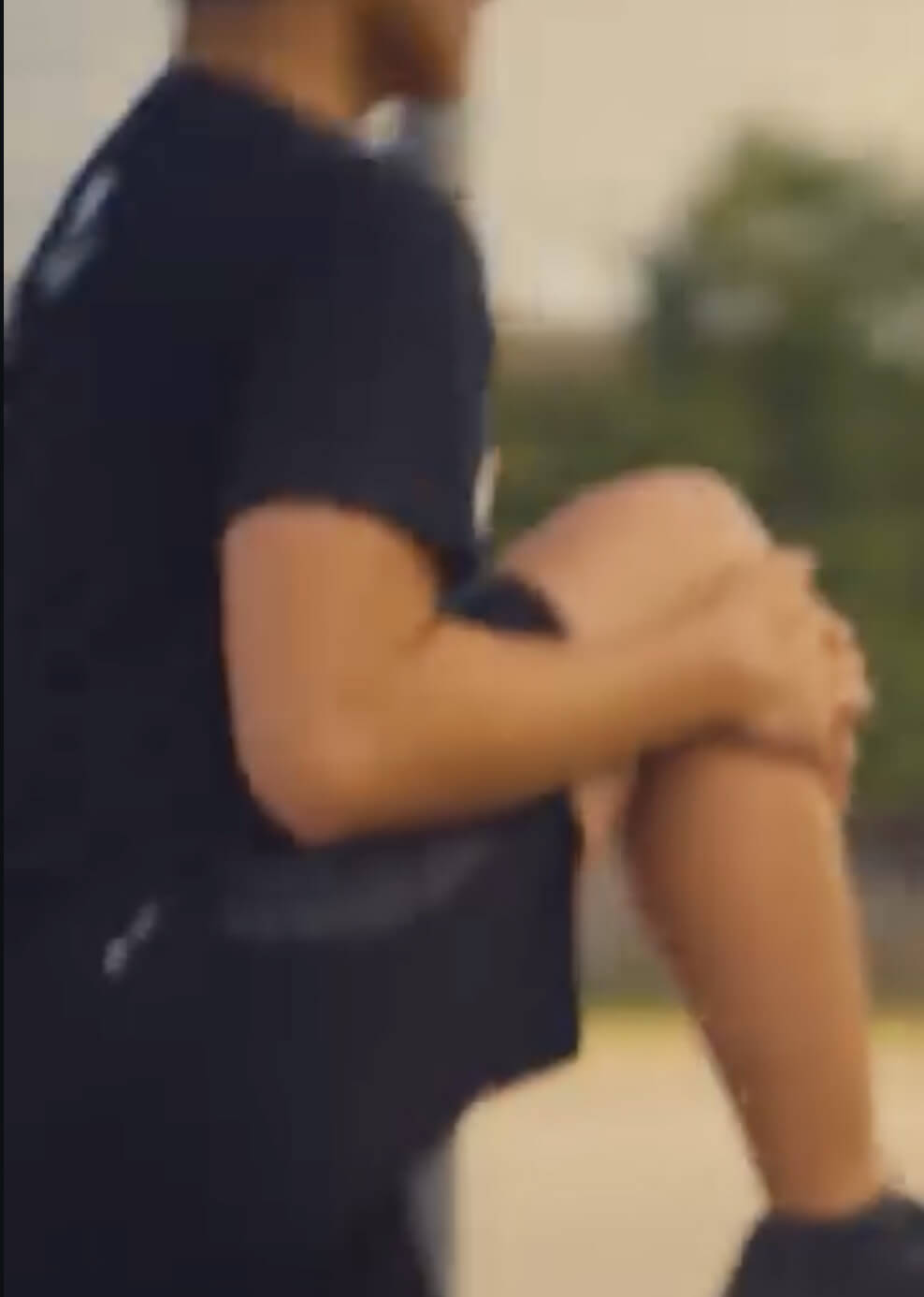 A person in a black shirt and shorts exercises outdoors, raising one knee toward their chest. The background is blurry, with hints of greenery and sky.