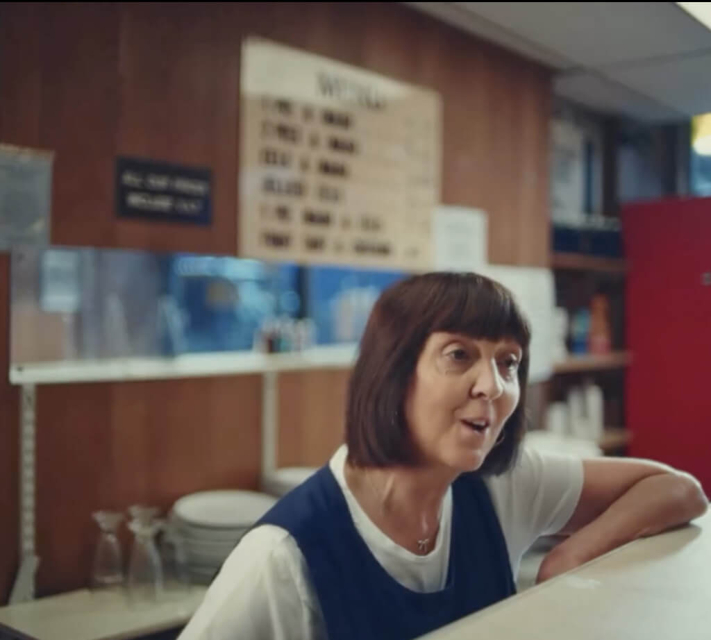 A woman with short dark hair, wearing a white shirt and blue apron, stands behind a counter in a diner or café. Shelves with dishes and a menu board are visible in the background.