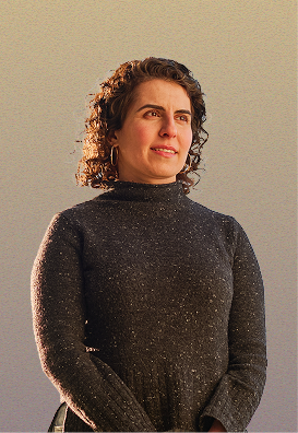 A woman with curly brown hair, wearing a dark gray long-sleeve turtleneck, stands against a neutral background, looking slightly to the side and smiling softly.