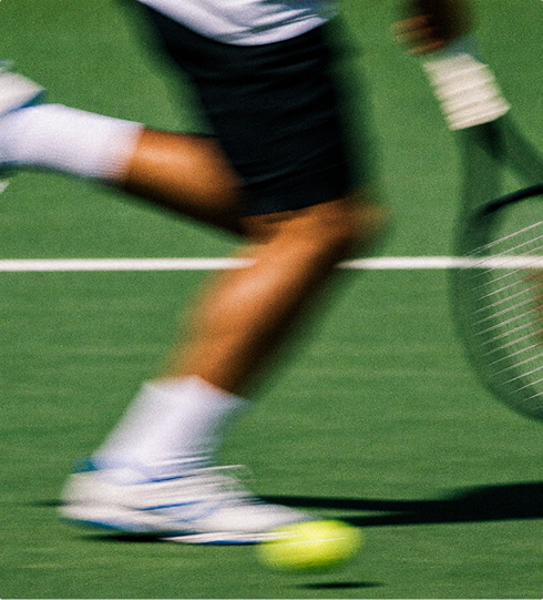 A blurred image of a tennis player running on a green court, with a racket in hand and a tennis ball on the ground, capturing the motion and speed of the sport.