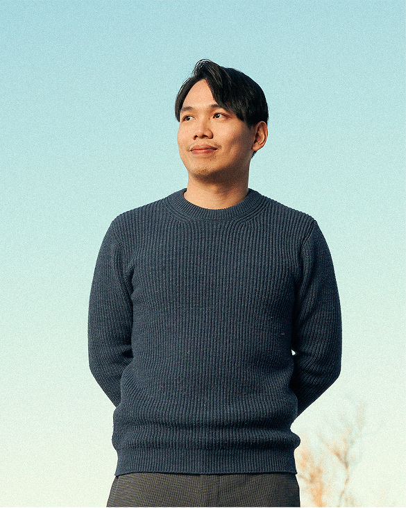 A person with short dark hair wearing a navy blue sweater stands outdoors against a clear blue sky, looking slightly to the side with a gentle smile and hands behind their back.