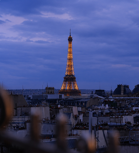 The Eiffel Tower glows with golden lights at dusk, rising above the rooftops of Paris under a cloudy blue sky.