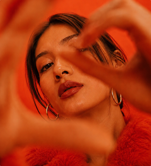 A woman with hoop earrings and red lipstick looks through her hands, which are forming a heart shape. She is wearing a red fuzzy top against a vibrant red background.