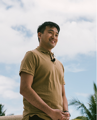 A man in a tan polo shirt smiles while standing outdoors against a partly cloudy sky, with palm tree leaves visible in the background.