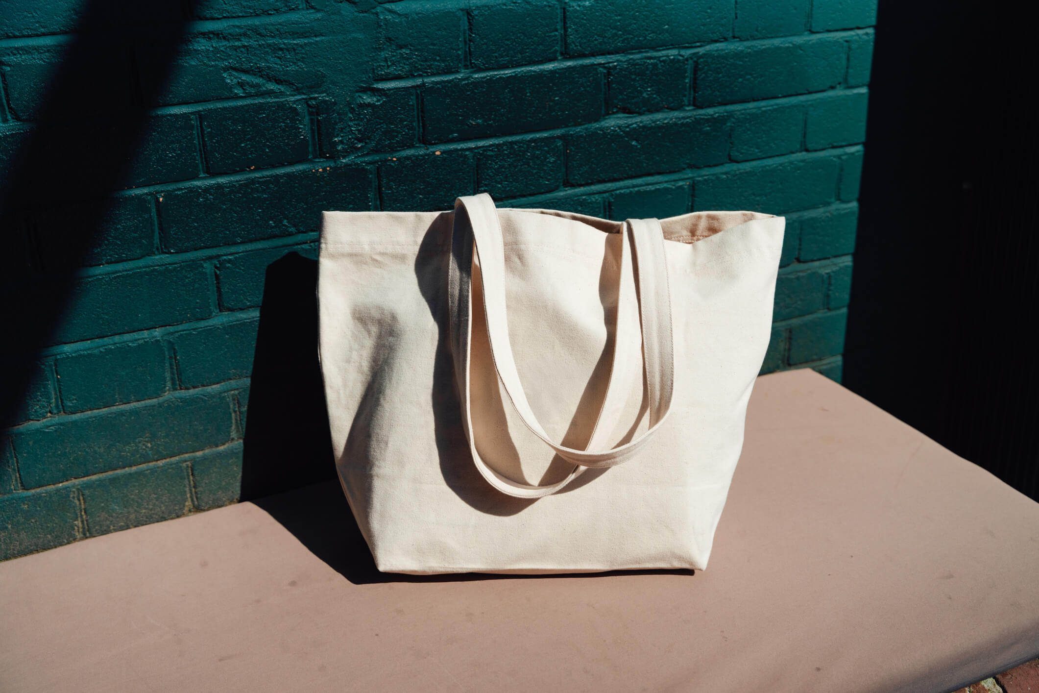 A plain beige canvas tote bag with two handles sits on a light brown surface in front of a green painted brick wall, with sunlight casting shadows across the scene.