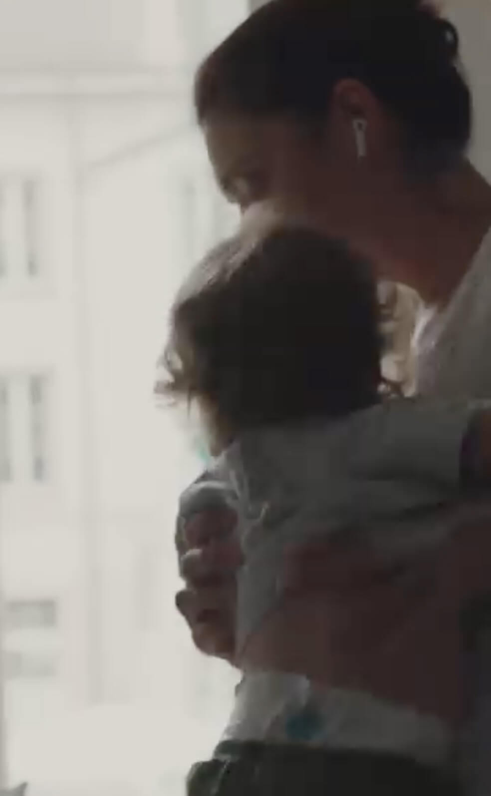 A woman wearing wireless earbuds holds a small child in her arms near a sunlit window, with blurred buildings visible outside.