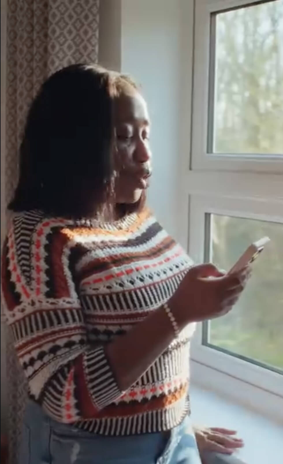 A woman wearing a colorful patterned sweater stands by a window, looking at her phone with sunlight illuminating her face. Trees and greenery are visible outside.