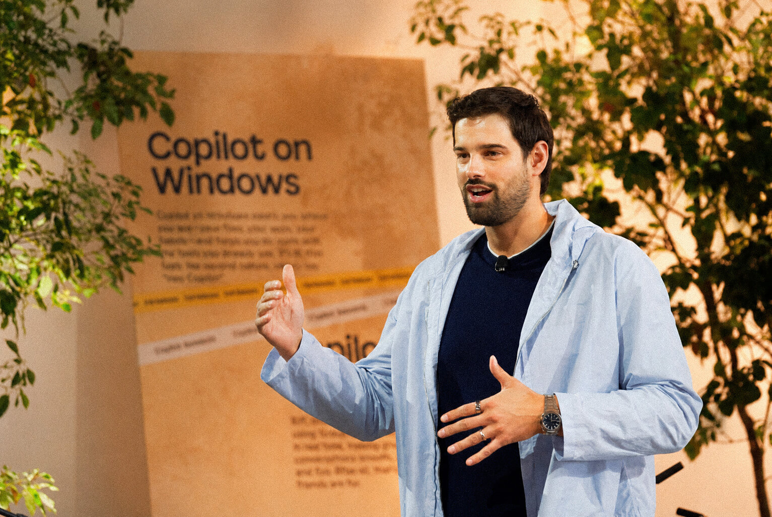 A man in a light blue jacket speaks and gestures during a presentation. Behind him is a sign that reads “Copilot on Windows” with text and greenery in the background.