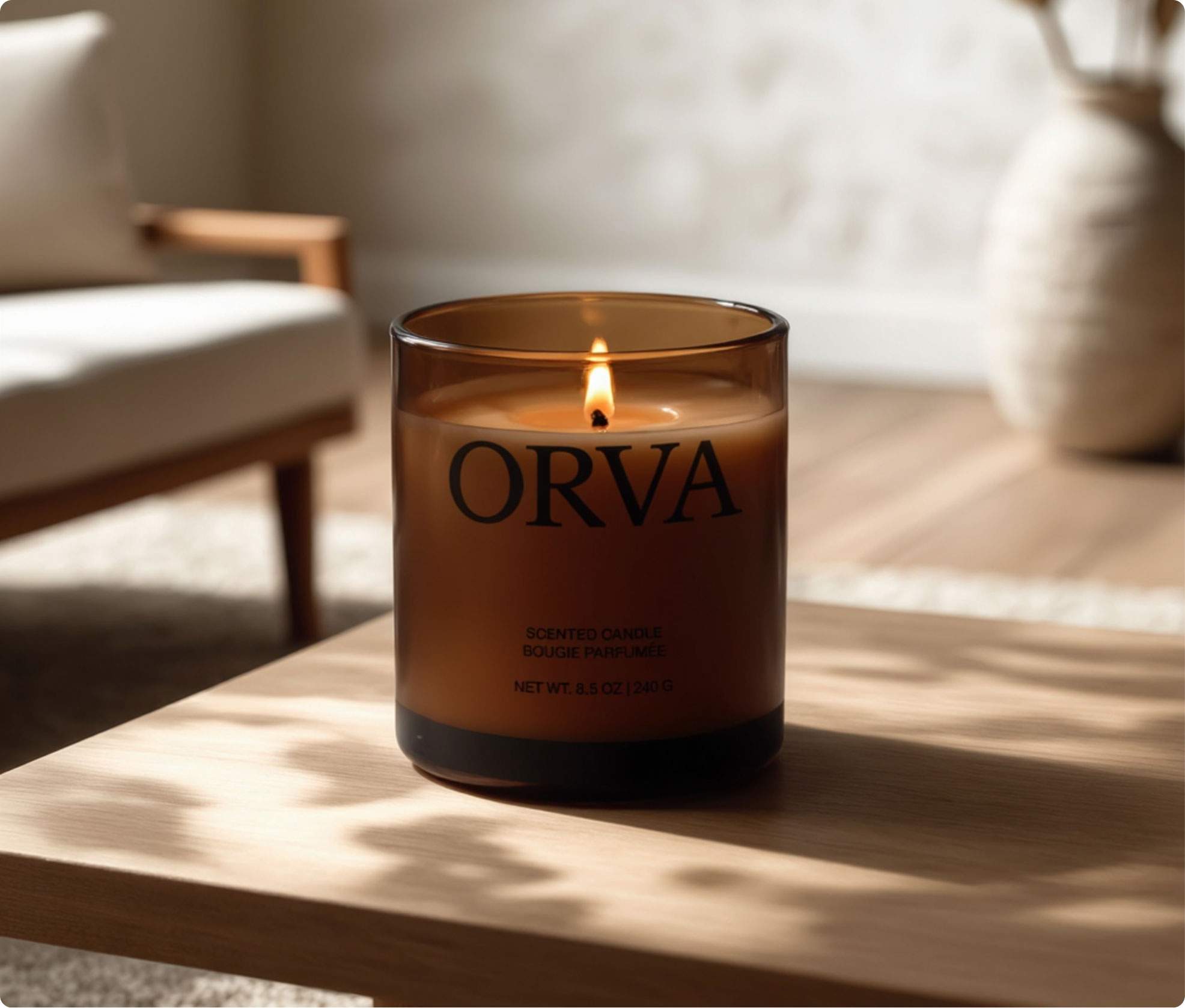 A lit amber glass candle labeled "ORVA" sits on a wooden table in a softly lit, cozy room with neutral decor and blurred furniture in the background.