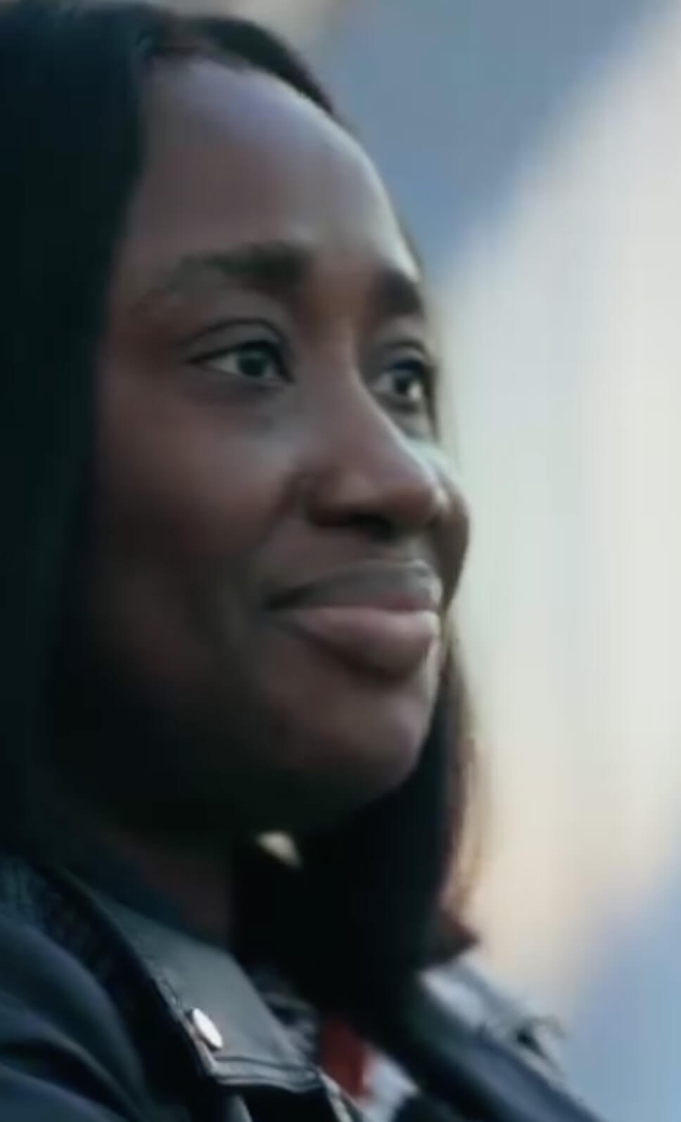 A woman with long, straight black hair smiles softly while looking to the side. She is wearing a dark jacket and is lit by natural light in an outdoor setting.