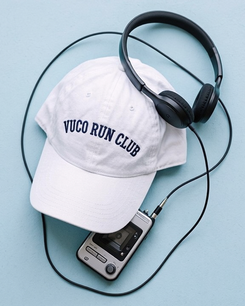 A white cap with "VUCO RUN CLUB" written on it sits on a light blue surface next to a black headset and a silver portable cassette player.