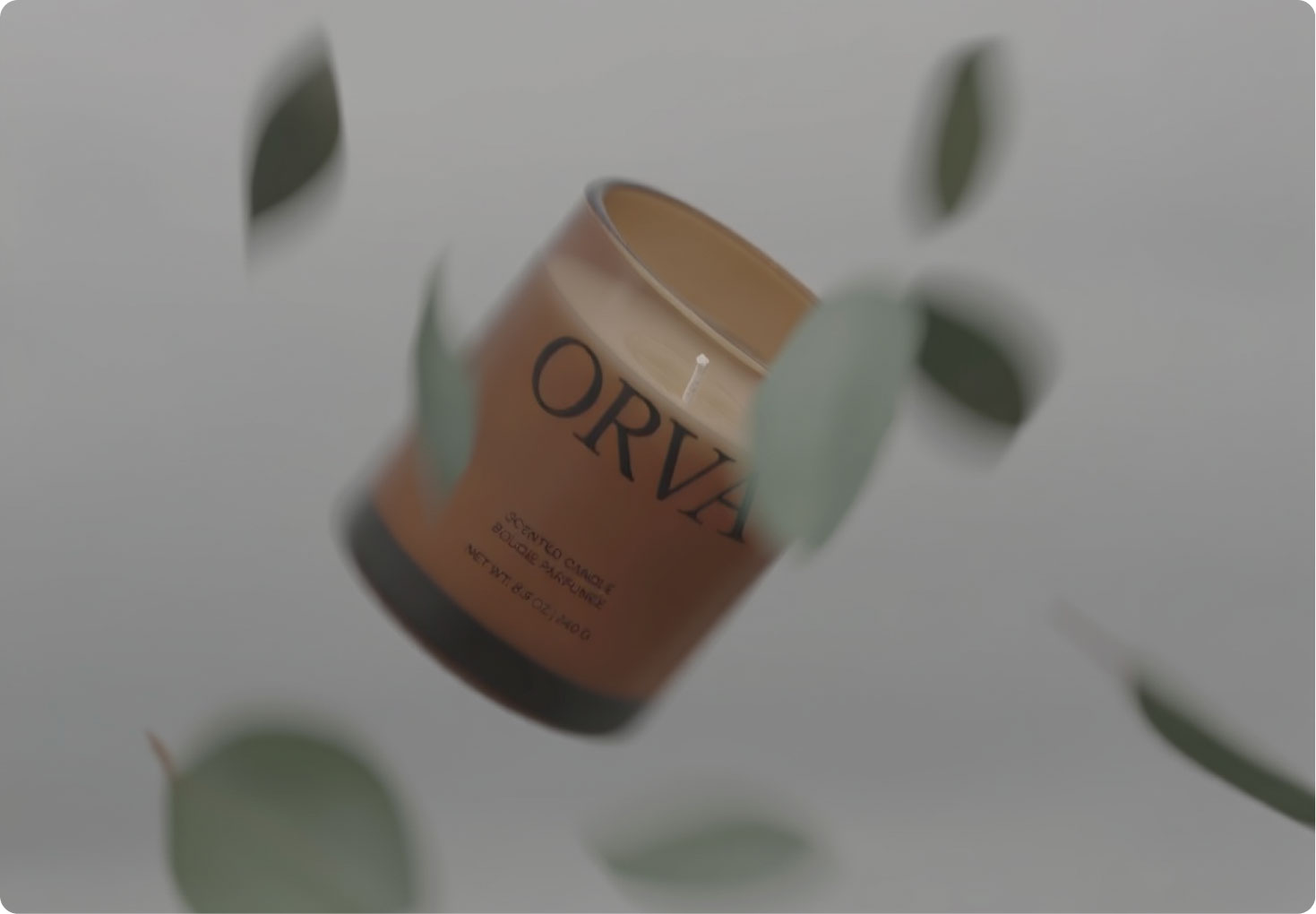 A brown candle jar labeled "ORVA" floats against a gray background, with green leaves blurred in the foreground, creating a soft, airy effect.