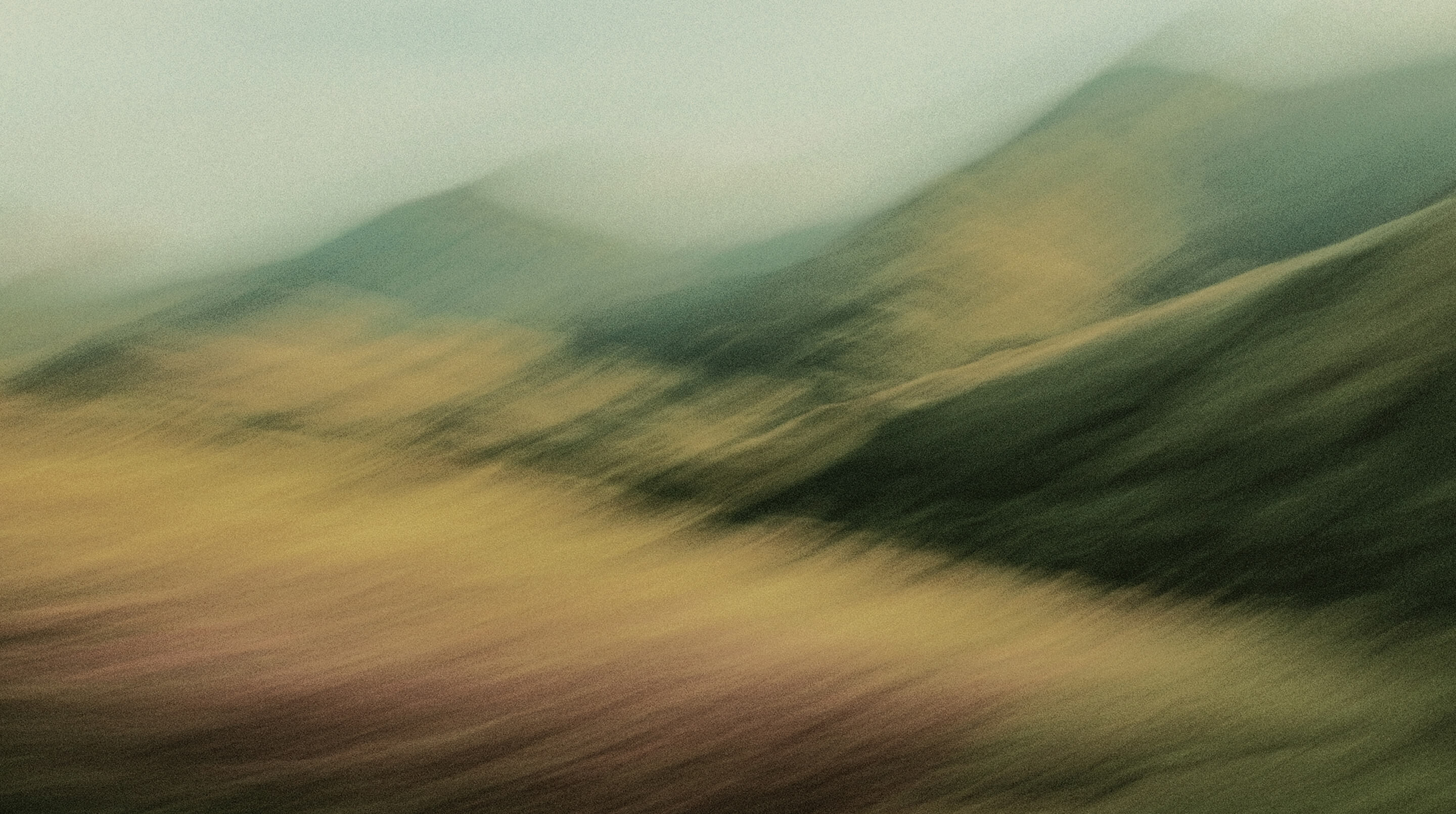 A blurred, abstract landscape featuring soft green and yellow hues, with hints of mountains and a field under a hazy sky. The image has a dreamy, painterly effect.