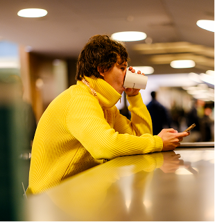 Person in a bright yellow sweater sitting at a counter, drinking from a white cup and looking at a smartphone, with a blurred indoor background.