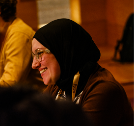 A woman wearing glasses and a black hijab smiles while sitting indoors. The background is softly lit and out of focus, creating a warm atmosphere.