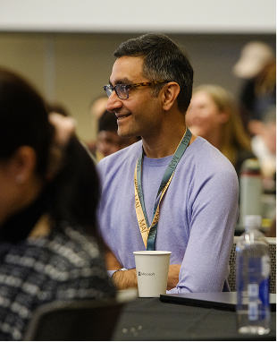 A man wearing glasses and a light purple shirt sits at a table with a lanyard around his neck, smiling and holding a paper cup. Other people are seated around him, and the setting appears to be a conference or seminar.