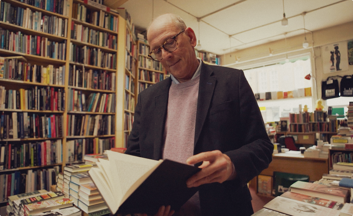 An older man wearing glasses and a blazer reads a book inside a cozy bookstore filled with shelves and stacks of books.