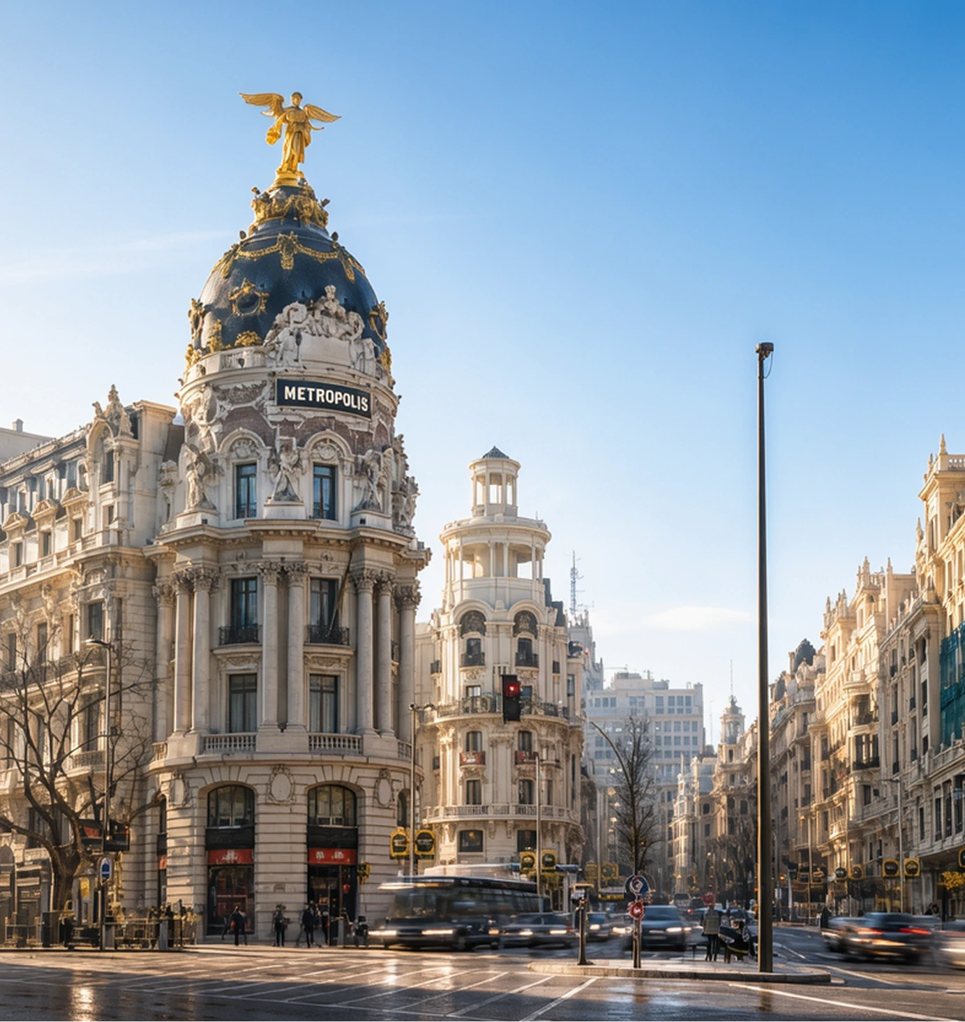 The Metropolis Building in Madrid stands at a busy intersection, topped with a golden winged statue. Surrounding ornate buildings line the street under a clear blue sky. Traffic and pedestrians are visible below.
